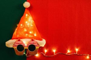 conceito de temporada de saudação chapéu de papai noel com luz de estrela e óculos que decoração com cupcake de natal em fundo vermelho e verde foto