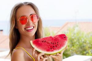 férias de verão baratas com tudo incluído. jovem mulher feliz com melancia e óculos de sol rosa no fundo da praia. fim de semana divertido de verão. linda garota com roupa de verão. foco seletivo. férias quentes foto