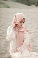 linda modelo feminina islâmica vestindo moda hijab, um vestido de noiva moderno para mulher muçulmana sentada na areia e na praia. retrato um modelo de menina asiática usando hijab se divertindo na praia com árvores foto