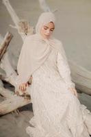linda modelo feminina islâmica vestindo moda hijab, um vestido de noiva moderno para mulher muçulmana sentada na areia e na praia. retrato um modelo de menina asiática usando hijab se divertindo na praia com árvores foto