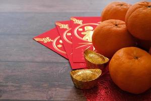 conceito de festival de ano novo chinês. tangerinas, envelopes vermelhos e lingotes de ouro em pano vermelho com fundo de madeira velho. caracteres chineses significam rico, rico, saudável e feliz. foto