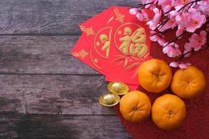 conceito de festival de ano novo chinês. tangerinas, envelopes vermelhos e lingotes de ouro em pano vermelho com fundo de madeira velho decorado com flor de ameixa. caractere chinês fu que significa sorte. foto