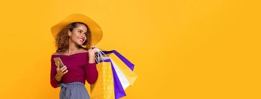 mulher de raça mista sorridente na moda com sacolas coloridas e celular isolado em fundo amarelo para conceito de venda de verão foto
