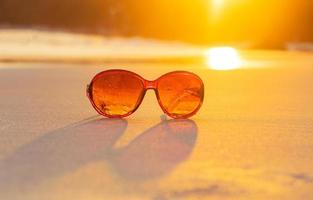 óculos de sol marrons na areia linda praia de verão e luz dourada do pôr do sol foto