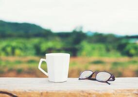 branco de xícara de café quente com óculos no banco com fundo de montanha verde natureza. foto