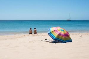 casal adulto sentado em uma praia olhando para o mar. visto de costas, perto de um guarda-sol colorido, sem pessoas ao redor, um iate ao longe. praia de sagres, algarve, portugal. foto