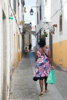mulher vista de costas andando em uma antiga rua em évora, portugal. vestindo um vestido colorido e carregando uma sacola plástica. um menino e um homem ao longe, vistos de costas. foto