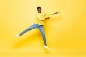 pulando o retrato de um jovem africano enérgico feliz abrindo as mãos para esvaziar o espaço de lado no fundo amarelo isolado do estúdio foto