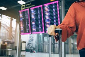 jovem mulher asiática em pé no aeroporto segurando a mala verificando a partida com o horário do voo em segundo plano. foto