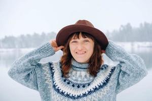 retrato de uma mulher sorridente em um chapéu marrom e suéter em um fundo de floresta coberta de neve e lago foto