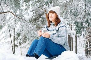 mulher com uma xícara de chá nas mãos sentada na floresta coberta de neve foto