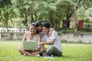 feliz família pai, mãe e filha sentada na grama e jogando laptop no parque ao ar livre foto