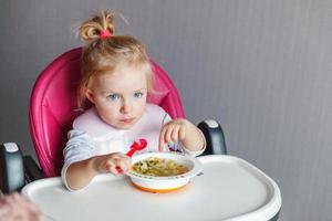 menina infantil com cara suja comendo sopa com colher na cadeira de bebê alta na cozinha em casa foto