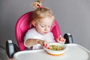 menina infantil com cara suja comendo sopa com colher na cadeira de bebê alta na cozinha em casa foto