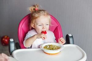 menina infantil com cara suja comendo sopa com colher na cadeira de bebê alta na cozinha em casa foto