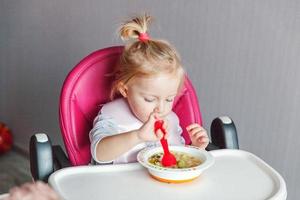 menina infantil com cara suja comendo sopa com colher na cadeira de bebê alta na cozinha em casa foto