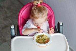 menina infantil com cara suja comendo sopa com colher na cadeira de bebê alta na cozinha em casa foto