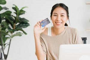 linda mulher asiática usando computador ou laptop para comprar compras on-line com cartão de crédito enquanto usa a camisola sentada na mesa na sala de estar em casa. mulher de estilo de vida no conceito de casa. foto