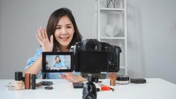 mulher asiática sorridente feliz ou blogueira de beleza com escova e câmera gravando vídeo e acenando com a mão em casa. beleza videoblog blogging pessoas conceito. foto