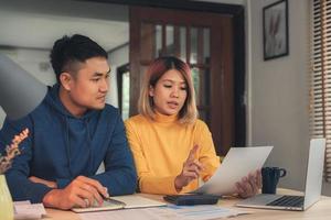 jovem casal asiático gerenciando finanças, revisando suas contas bancárias usando computador portátil e calculadora em casa moderna. mulher e homem fazendo papelada juntos, pagando impostos on-line no notebook. foto