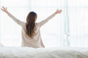 vista traseira da bela jovem asiática feliz acordando de manhã, sentado na cama, esticando-se no quarto aconchegante, olhando pela janela. mulher engraçada depois de acordar. ela está esticando e sorrindo. foto