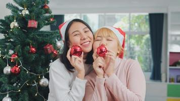 casal de lésbicas asiáticos celebra o festival de natal. lgbtq feminino adolescente usar chapéu de natal relaxar feliz jogar bola de natal desfrutar de férias de inverno de natal juntos na sala de estar em casa. foto