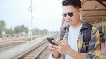 homem de mochila asiático na estação de trem, jovem mochileiro de turista masculino usando o celular olhando e mapas de direção em sua jornada na tailândia. homens de estilos de vida relaxam e viajam o conceito. foto