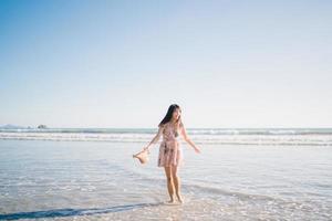 jovem mulher asiática caminhando na praia. linda fêmea feliz relaxe caminhando na praia perto do mar ao pôr do sol à noite. mulheres de estilo de vida viajam no conceito de praia. foto