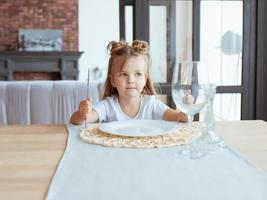 retrato de menina adorável com fome em camiseta branca, sentado à mesa com garfo, faca e prato vazio no interior do sotão foto
