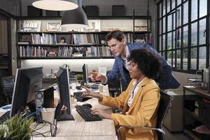 jovem trabalhadora afro-americana senta-se, trabalha com computador, brainstorming, conversando e discutindo com colega caucasiano e parceria sobre empregos de negócios na empresa de escritório do espaço de trabalho. foto