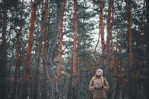 garota atraente em roupas de inverno ee um copo de bebida quente está andando na floresta. foto