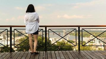 vista traseira de uma jovem de camisa branca, um viajante morena olha para a paisagem urbana em um dia ensolarado de uma colina alta com um deck de observação para turistas. vintage foto