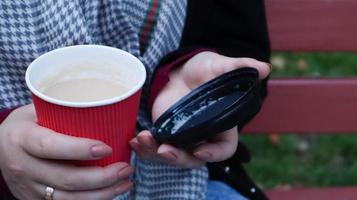 mãos femininas estão segurando o café da manhã para viagem e fechando um copo de papel com tampa enquanto está sentado em um banco do parque, close-up das mãos. foto