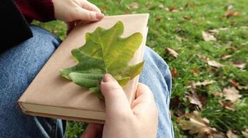 uma mulher segura um livro fechado no colo com uma folha de carvalho caída close-up em um parque em um dia ensolarado de outono quente. o conceito de relaxamento, leitura e relaxamento sozinho. foto