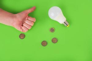 lâmpada de poupança de energia, sinal de aprovação à mão e moedas de euro em um fundo verde. vista do topo. conceito de economia de energia. lugar para texto foto