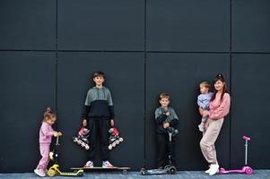 jovem mãe elegante com quatro filhos ao ar livre contra a parede moderna preta. família de esportes passa o tempo livre ao ar livre com patinetes e patins. foto