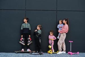 jovem mãe elegante com quatro filhos ao ar livre contra a parede moderna preta. família de esportes passa o tempo livre ao ar livre com patinetes e patins. foto