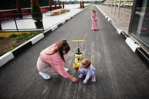 jovem mãe elegante com duas meninas ao ar livre. família de esportes passa o tempo livre ao ar livre com scooters. pintado com giz no asfalto. foto