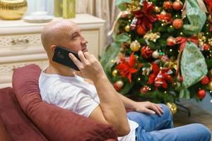 jovem careca feliz senta-se na frente da árvore de natal decorada em seu apartamento foto