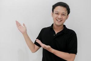 feliz e sorriso de jovem asiático com ponto de mão no espaço vazio. homem indonésia usa camisa preta isolado fundo cinza foto