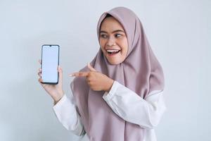 jovem islão asiático usando lenço na cabeça está apontando o dedo na tela branca do telefone com sorriso e sentimento feliz. mulher indonésia em fundo cinza foto
