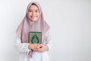 jovem islão asiático vestindo lenço na cabeça está segurando o alcorão sagrado na mão com sorriso e cara feliz. mulher indonésia em fundo cinza foto