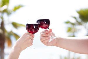 mãos de homem e mulher segurando copos de vinho tinto de suco de cereja, brindando, em fundo de verão tropical. conceito de férias de viagem. copie o espaço. foto