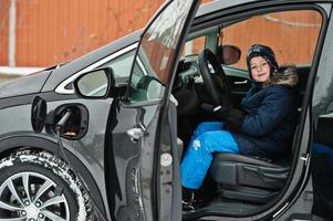 menino senta-se no carro e carrega o veículo elétrico no quintal da casa no inverno. foto