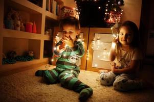 duas irmãs olhando na árvore de natal com guirlandas brilhantes na noite em casa. foto