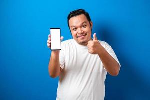 satisfeito feliz jovem asiático gordo em camiseta branca sorrindo para a câmera enquanto segura o smartphone apontando para a tela do celular foto