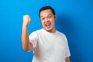 retrato de um homem asiático gordo engraçado em t-shirt branca sorrindo e dançando alegremente, alegre expressando comemorando boas notícias vitória ganhando gesto de sucesso contra fundo azul foto