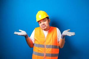 jovem trabalhador asiático gordo bonito vestindo uniforme de colete de segurança laranja e expressão sem noção e confusa de capacete com braços e mãos levantadas foto