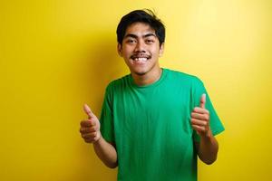 jovem asiático bonito vestindo camiseta verde de pé sobre fundo amarelo isolado fazendo feliz polegares para cima foto