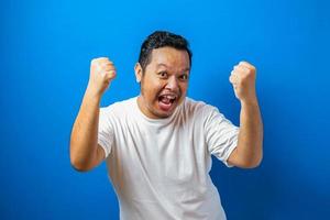 retrato de um homem asiático gordo engraçado em t-shirt branca sorrindo e dançando alegremente, alegre expressando comemorando boas notícias vitória ganhando gesto de sucesso contra fundo azul foto